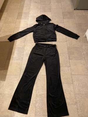 Svart mjukisset från Gina Tricot - Svarta mjukisbyxor och hoodie från Gina Tricot i mjukt och glansigt material. Byxorna har resår i midjan med snörning och vida ben. Perfekta för en chill och avslappnad stil. Matcha med hoodie för en komplett look.