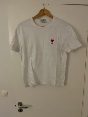 Vit t-shirt från Ami med rött hjärta - Säljer en clean vit t-shirt från Ami Alexandre Mattiussi med ett broderat rött hjärta och bokstaven A på bröstet. Klassisk rund hals och korta ärmar. Materialet är mjuk bomull som känns skönt mot huden. Perfekt för dig som gillar stilrena plagg med en twist.