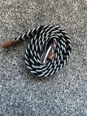 Svartvit flätad tygbälte - Säljer ett svartvitt flätat bälte med silverfärgat spänne och bruna detaljer i ändarna. Bältet är gjort i ett mjukt och flexibelt tygmaterial och har ett randigt mönster som ger en cool och modern look. Perfekt för att lyfta vilken outfit som helst.