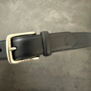 Svart skärp från Polo Ralph Lauren - Har ett i storlek 110cm och ett exakt likadant i 100cm. Säljer ett stilrent svart skärp från Polo Ralph Lauren. Skärpet har en klassisk silverfärgad metallspänne och den ikoniska Polo-loggan präglad på lädret. 