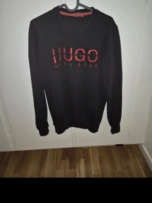 Svart tröja från Hugo Boss - Säljer en svart långärmad tröja från Hugo Boss med rött tryck på bröstet. Tröjan har en klassisk rund hals och är tillverkad i mjukt material som passar perfekt för en chill och stilren look. Perfekt för dig som gillar snygga logotröjor.