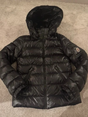 Svart Moncler pufferjacka med huva - Säljer en svart pufferjacka från Moncler med glansig finish och huva. Jackan har två dragkedjefickor framtill, Moncler-logga på ärmen och quiltad design. Perfekt för kalla dagar och riktigt snygg streetstil.