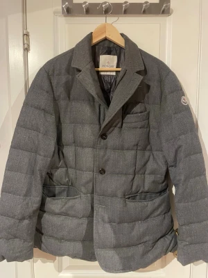 Grå quiltad dunjacka från Moncler - Säljer en grå quiltad dunjacka från Moncler med kavajkrage och knäppning framtill. Jackan har två fickor fram och Moncler-logga på ärmen. Stilsäker modell som kombinerar klassisk kavajlook med värmande dunfoder. Perfekt för kalla dagar när du vill vara både varm och snygg.