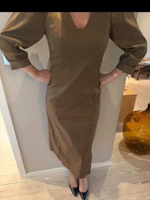 Brun långklänning från Nümph - Säljer en snygg brun långklänning från Nümph i storlek 36. Klänningen har v-ringning, trekvartsärmar med puff och markerade axlar. Materialet känns som bomull och modellen är figurnära med slits bak. Perfekt för dig som gillar stilrena och trendiga plagg.