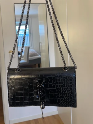 Svart axelväska från Yves Saint Laurent - Säljer en svart axelväska från Saint Laurent med croco-mönstrad yta i skinn och kedjeremmar i metall. Väskan har YSL-logga framtill och en snygg detalj med hängande dragkedja. Insidan är fodrad i rött tyg och har ett innerfack. Perfekt för dig som vill ha en lyxig och stilren look.