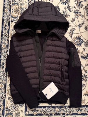 Svart Moncler Cardigan - Säljer en svart pufferjacka från Moncler med stickade ärmar och stor huva. Jackan har dragkedja framtill, ribbade muddar och en snygg Moncler-logga på ärmen. Perfekt för dig som vill ha en stilren och varm jacka med exklusiv känsla.