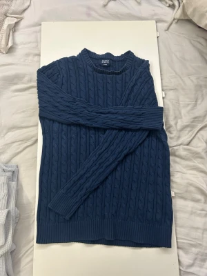 Mörkblå kabelstickad tröja Iconic Spirit - Mysig mörkblå kabelstickad tröja från Iconic Spirit i storlek L. Men passar även M. Och nästan S Tröjan har rund hals, långa ärmar och breda ribbade muddar vid ärmslut och nederkant. Perfekt för kyliga dagar och enkel att matcha med jeans.