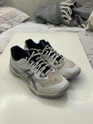 Vita ASICS sneakers med grå detaljer - Säljer ett par vita ASICS sneakers med grå och svarta detaljer. Skorna har snörning, rund tå och platt sula. Ovandelen är i syntetmaterial med meshpaneler för extra ventilation. Perfekta för träning eller sport. Snyggt ASICS-logga på sidan.