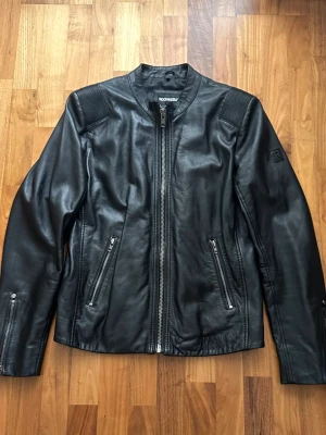 Svart skinnjacka från Rockandblue - Säljer en svart skinnjacka från Rockandblue med coola detaljer på axlarna och två snedställda fickor med dragkedja. Jackan har en rak passform, rund krage och grov dragkedja framtill. Perfekt för dig som vill ha en stilren och edgy look.