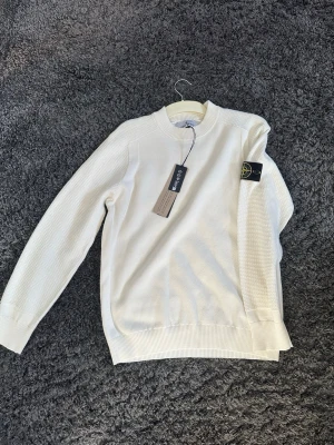 Vit stickad tröja från Stone Island - Säljer en clean vit stickad tröja från Stone Island med klassisk patch på ärmen. Tröjan har rund hals, ribbade muddar och struktur på ärmar och axlar. BNWT.