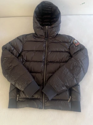 Svart pufferjacka från Parajumpers - Säljer en svart pufferjacka från Parajumpers med huva och emblem på ärmen. Jackan har quiltade partier på armbågarna, två fickor med dragkedja och breda muddar vid ärmslut och nederkant. Perfekt för kalla dagar och riktigt snygg streetstil.