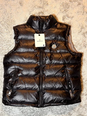 Svart dunväst från Moncler 4/L - Svart dunväst från Moncler med glansig finish och quiltad design. Västen har två fickor med dragkedja, hög krage och klassisk Moncler-logga på bröstet. . Dragkedja framtill och coolt innerfoder med serietidningsmotiv. Skriv vid frågor storlek 4L