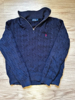 Marinblå stickad tröja Polo Ralph Lauren - Kabelstickad marinblå tröja från Polo Ralph Lauren med halv dragkedja och klassisk röd logga på bröstet. Tröjan har ribbade muddar och hög krage, perfekt för kyliga dagar. Snygg och tidlös design som funkar till allt. Längden på tröjan är cirka 60 cm och axelängden är cirka 48 cm