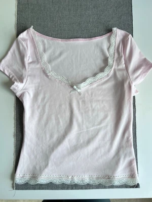 Ljusrosa topp med spets och rosett, storlek S - Ljusrosa topp med korta ärmar, v-ringad hals och vita spetskanter. Framtill sitter en liten vit rosett 🎀🤍 Materialet är mjukt och väldigt stretchigt. - Köper du innan kl 16:00 kan jag posta samma dag!
