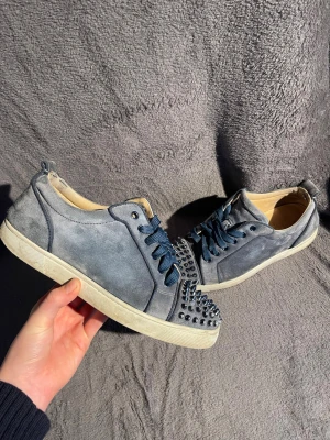 Grå mocka sneakers med nitar - Säljer ett par grå sneakers i mocka från Christian Louboutin med blå snören och ikonisk röd sula. Skorna har coola metallnitar över tån och en klassisk låg siluett. Perfekta för dig som vill sticka ut med detaljer och exklusiv känsla.