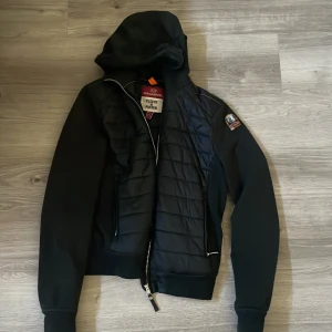 Parajumpers CAELIE Girl  - Svart jacka från Parajumpers i kombinerat fleece- och puffer-material. Jackan har huva, quiltad front, dragkedja och klassisk PJS-patch på ärmen. Litet hål som säkert går att laga men jag är inte så sykunnig. Storlek 16 år