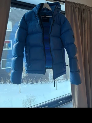 Blå Peak Performance dunjacka - Säljer en blå dunjacka från Peak Performance med huva och dragkedja framtill. Jackan har en puffad design och är perfekt för kalla dagar. Färgen är klarblå och materialet är syntet med dunfyllning. Nypris 4999