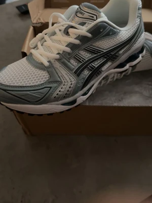 Asics GEL-Kayano 14 sneakers vit/silver - Säljer ett par Asics GEL-Kayano 14 sneakers i vitt och silver med mesh-överdel och detaljer i syntet. Skorna har klassisk snörning, sportig vibe och en chunky sula med GEL-dämpning för extra komfort. Perfekta för dig som gillar retrostil och streetwear.