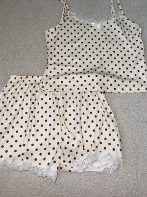 Prickig väst med spetsdetaljer - Söt väst i vit med svarta prickar och tunna axelband. Dekorerad med vit spets och liten rosett framtill. Materialet känns mjukt och lätt, perfekt för en chill look. Matchar enkelt med andra plagg för en lekfull vibe.