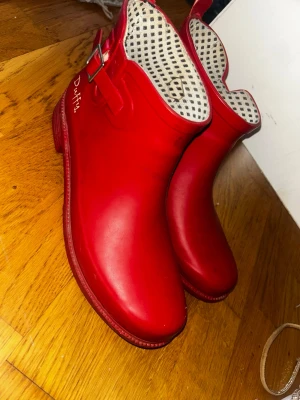 Röda korta stövlar från Duffy - Snygga röda korta stövlar från Duffy med spänndetalj på sidan och svartvitt prickigt innerfoder. Skorna har rund tå och platt sula, perfekta för regniga dagar. Tillverkade i syntetmaterial som håller fötterna torra.