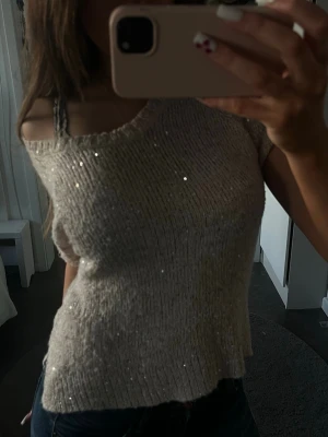 Beige glittrig stickad topp - Säljer en beige stickad topp med glittriga paljettdetaljer. Toppen har en lös passform och är kortärmad med en bred, något offshoulder ringning. 