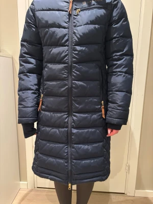 Mörkblå ridjacka/ridkappa från Jacson - Säljer en lång, mörkblå varm ridjacka/ridkappa från Jacson med huva och dragkedja framtill. Jackan har snygga gulddetaljer på dragkedjor och knappar bak, samt praktiska fickor.  Perfekt för kalla dagar i stallet. Mkt bra skick! Nypris 1400kr.