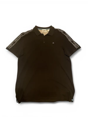 Svart Calvin Klein piké med logga - Svart pikétröja från Calvin Klein i mjuk pima bomull. Klassisk krage, korta ärmar och vita logoband längs axlarna. Liten CK-brodyr på bröstet och knappslå framtill. Perfekt för en clean och sportig look.