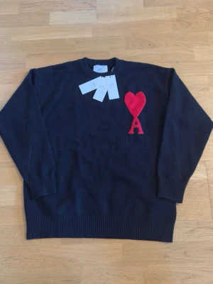 Svart stickad tröja från Ami - Svart stickad tröja från Ami Alexandre Mattiussi med röd hjärtlogga och bokstaven A på bröstet. Klassisk rund halsringning och ribbade muddar vid ärmar och nederkant. Perfekt för dig som gillar stilrena plagg med en twist.