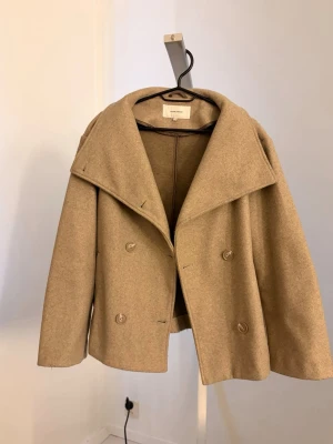 Beige kort kappa  - En kort beige kappa med dubbelknäppning från Bubbleroom. 
