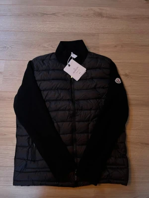 Moncler cardigan  - Svart Cardigan från Moncler med quiltad front och stickade ärmar. Jackan har dragkedja, ståkrage och Moncler-logga på ärmen. Materialet är en mix av polyester och stickad textil, perfekt för kyliga dagar. Snygg och stilren design med coola detaljer. Helt ny! Aldrig använd!
