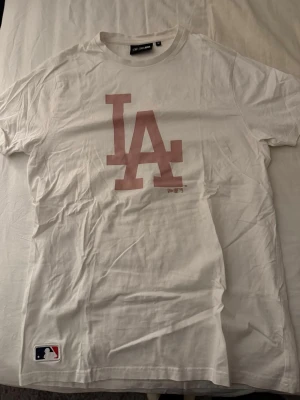 Vit LA t-shirt  - Säljer en vit t-shirt med stort LA-tryck i rosa på bröstet och MLB-logga nertill. Klassisk rund hals och korta ärmar. Perfekt för dig som gillar sportig streetstyle och vill ha en clean look.
