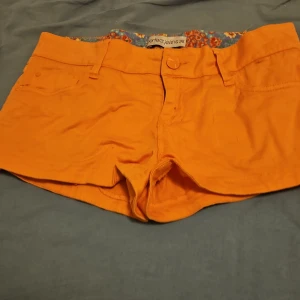 Orange shorts från Perfect Jeans - Snygga orange shorts från Perfect Jeans med klassisk femficksdesign och knappstängning. Insidan av linningen har ett färgglatt blommigt tyg i lila, rött och grönt. Perfekta för varma dagar och ger en riktigt poppig look.