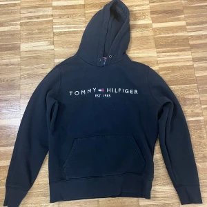 Svart hoodie från Tommy Hilfiger - Svart hoodie från Tommy Hilfiger med klassisk logga och texten 'EST. 1985' på bröstet. Tröjan har huva med dragsko och en stor magficka. Perfekt för dig som gillar en clean och stilren look.