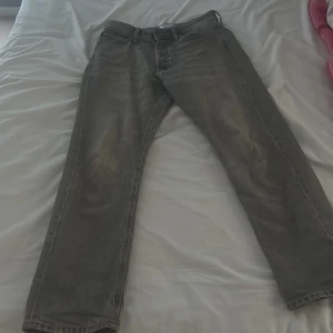 Gråa relaxed jeans från Jack & Jones - Gråa jeans från Jack & Jones i modellen Relaxed/Chris. Jeansen har en avslappnad passform med raka ben och klassisk femficksdesign. Tillverkade i mjukt jeanstyg med snygg tvättad look och bälteshällor i midjan.