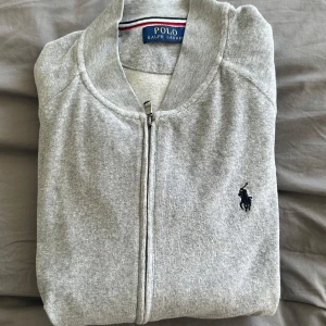 Grå zip-kofta från Polo Ralph Lauren - Grå kofta med dragkedja från Polo Ralph Lauren. Mjuk bomullskvalitet och långärmad modell som passar perfekt till chill dagar. Ganska stor i storleken 