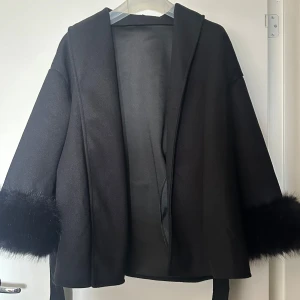 Svart kappa med pälsdetaljer och bälte - Säljer en svart kappa med bred krage och snygga pälsdetaljer vid ärmsluten. Jackan har ett omlott-utseende och knyts med bälte i midjan. Modellen är loose fit och har långärmade ärmar. Perfekt för dig som vill ha en stilren och trendig look.