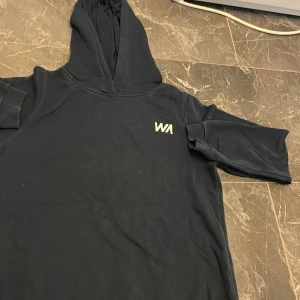 Svart hoodie från Warp med tryck - Svart hoodie från Warp med vit logga på bröstet och stort tryck med bladmönster och Warp-text på ryggen. Klassisk huva och långa ärmar, tillverkad i mjuk bomullsmix. Perfekt för chill dagar eller när du vill ha en enkel streetstyle-look.