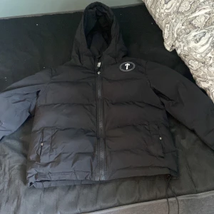 Svart pufferjacka från Trapstar XS - Svart pufferjacka från Trapstar med huva och dragkedja framtill. Jackan har Trapstar-logga på bröstet och stor broderad text på ryggen. Fylld för extra värme och har två fickor med knappstängning. Perfekt för kalla dagar och streetwear-stil.