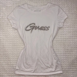 Vit t-shirt med glitterlogo från Guess - Vit t-shirt från Guess i bomull med korta ärmar och figurnära passform. Stort Guess-tryck framtill i glittriga silverpaljetter som ger en extra snygg touch. Perfekt för dig som gillar att sticka ut med stilrena detaljer.