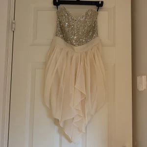 Champagnefärgad paljettklänning - Superfin kort klänning i champagnefärg med glittrig paljettbyst och draperad kjol. Axelbandslös modell med v-ringning och volangliknande lager i kjolen. Perfekt för dig som vill sticka ut med en elegant och glamorös look.