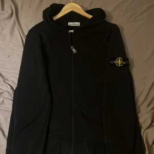 Svart hoodie från Stone Island - Svart hoodie från Stone Island med dragkedja framtill och klassisk logga på vänster ärm. Tröjan har huva och ribbade muddar. Perfekt för dig som gillar streetwear och vill ha en stilren look.