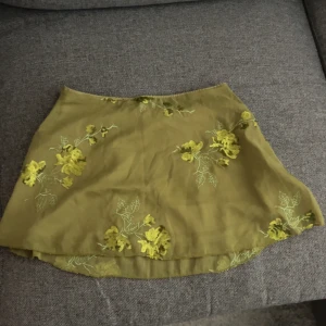 Olivgrön blommig kortkjol med paljetter - Aldrig andvänts! Supersöt olivgrön kortkjol med broderade blommor och glittriga paljettdetaljer. Kjolen har en lätt A-linjeform och är perfekt för dig som gillar unika detaljer och blommigt mönster. Materialet känns lätt och flowy.