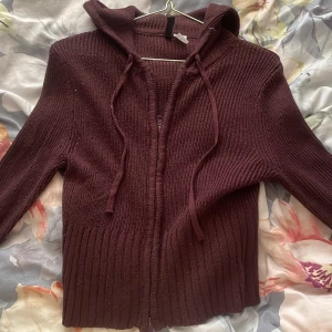 Vinröd stickad hoodie med dragkedja - Mysig vinröd stickad hoodie från H&M med dragkedja framtill och snörning i huvan. Tröjan har ribbade muddar och lång ärm, perfekt för kyliga dagar. Snygg och enkel att matcha med jeans eller kjol.
