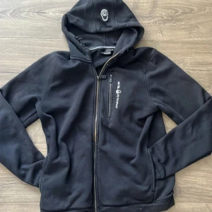 Svart hoodie från Sail Racing med dragkedja - Svart hoodie från Sail Racing med broderad logga på bröstet och huvan. Tröjan har hel dragkedja framtill, bröstficka med dragkedja och mjukt fleecefoder på insidan. Perfekt för kyliga dagar och snygg till jeans eller träningsbyxor.