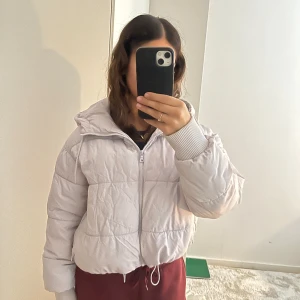 Ljuslila pufferjacka från Monki - Säljer en ljuslila pufferjacka från Monki i storlek S. Jackan har huva med snörning, dragkedja framtill och breda ribbade muddar vid ärmsluten. Quiltad design och croppad passform ger en trendig look. Perfekt för kyliga dagar!