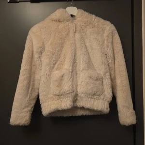 Beige teddyjacka med öron på huvan - Supermjuk och fluffig beige teddyjacka med dragkedja framtill, två stora fickor och huva med söta öron. Perfekt för dig som vill ha en mysig och trendig look. Långärmad och i oversized modell för extra komfort.