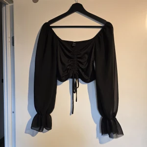 Svart croppad blus med snörning - Snygg svart blus med croppad passform och långa, transparenta ärmar i chiffongliknande material. Toppen har snörning framtill. Den var trasig i kanten när jag fick den.