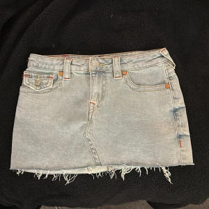 True Religion Sadie jeanskjol ljusblå - Ljusblå jeanskjol från True Religion, modell Sadie Mini Skirt. Kjolen har rå, fransig nederkant, klassiska fickor fram och bak med färgglada broderier och logoknappar. 