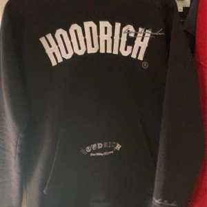 Svart HOODRICH hoodie med tryck - Svart hoodie från HOODRICH med stor vit logga på bröstet och mindre tryck på fickan och huvan. Klassisk känguruficka och huva med tryck. Perfekt streetstyle-plagg med lång ärm och relaxed vibe.