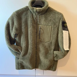 Olivgrön teddyjacka från Stone Island - Säljer en olivgrön teddyjacka från Stone Island med hög krage och dragkedja framtill. Jackan har en bröstficka med dragkedja och den klassiska Stone Island-loggan på ärmen. Materialet är mjuk fleece som ger en skön vibe och jackan har en loose passform.